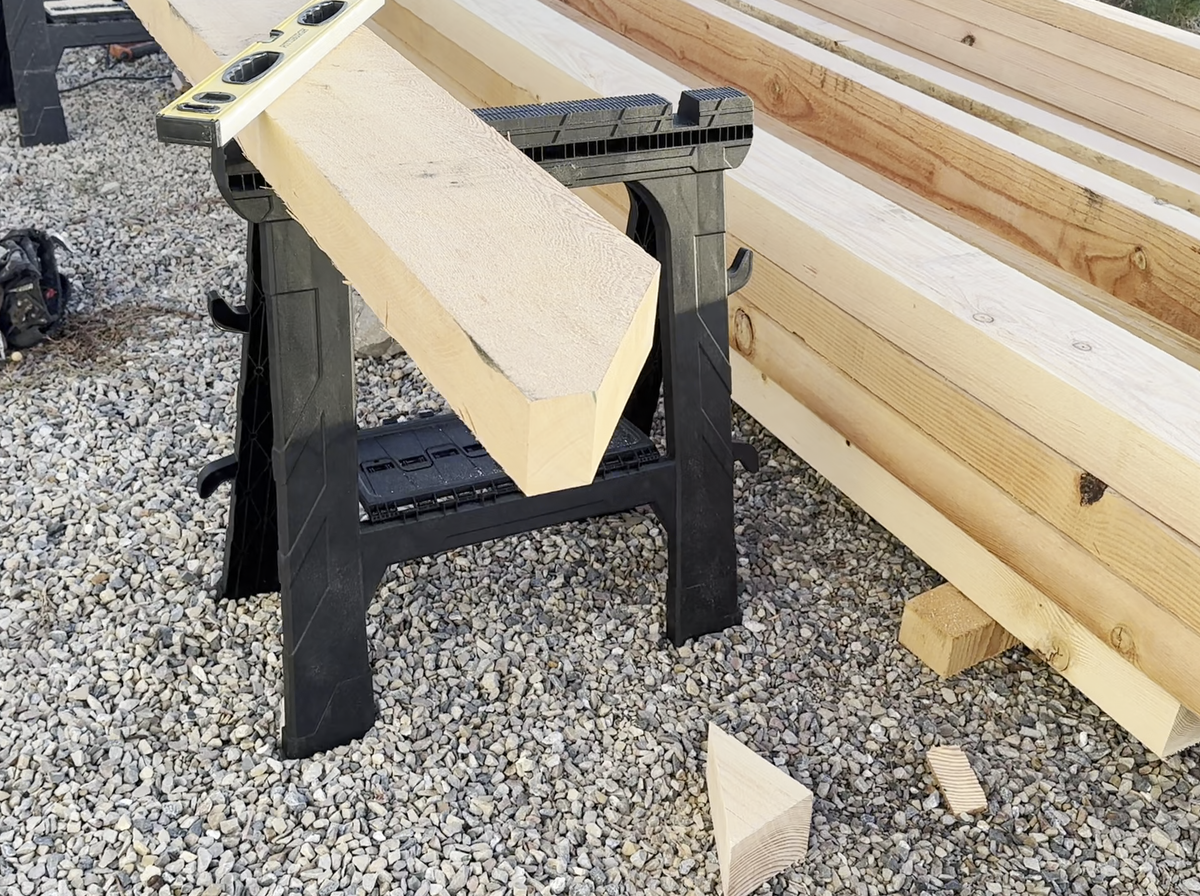 A Douglas fir timber with its end cut to shape, resting on a sawhorse beside a stack of cut timbers, offcuts on the gravel