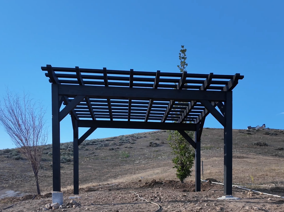 Slate 12×16 timber pergola with straight knee braces standing on a dry hillside lot in Lehi, Utah, under a clear blue sky