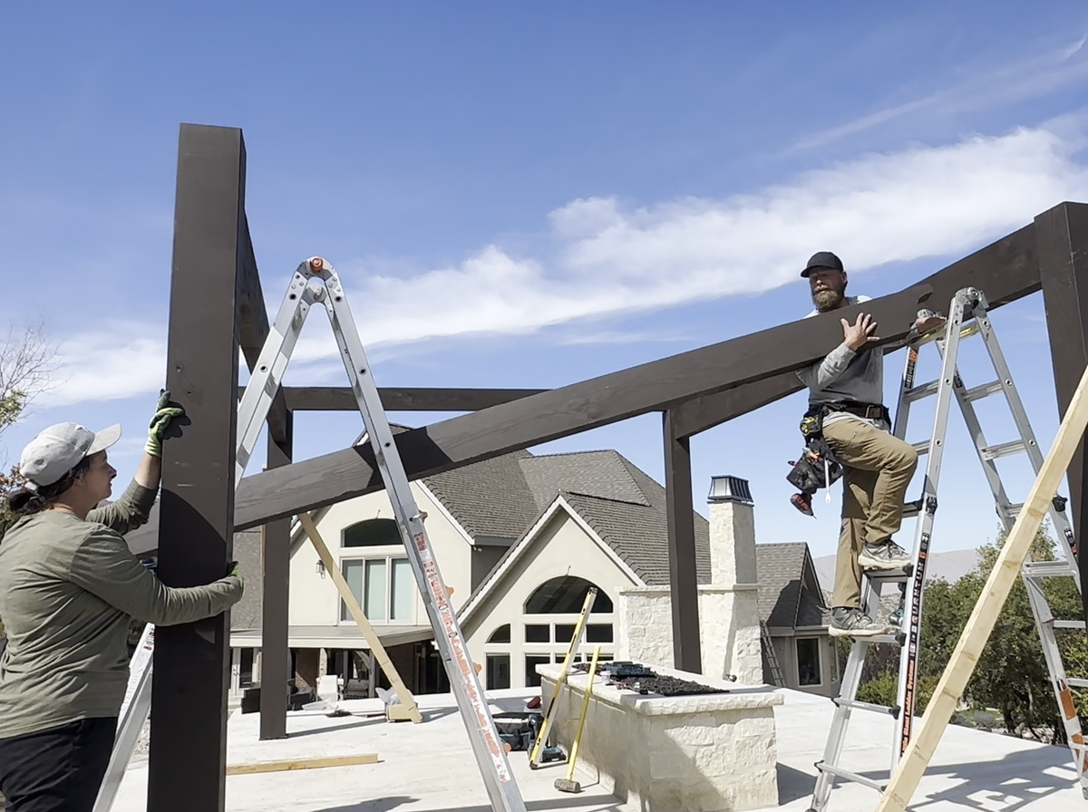 Two crew lifting a Cordovan Brown beam onto braced timber posts from a ladder during the build in Elk Ridge, Utah