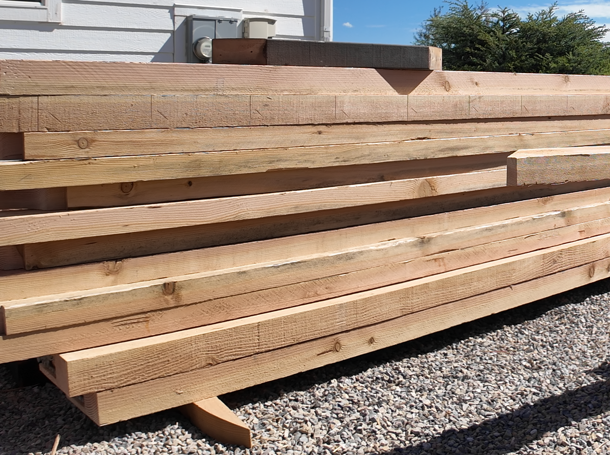 Rough-sawn Douglas fir timbers for an 18×18 pergola stacked before staining, the top one notched and the next marked out in pencil